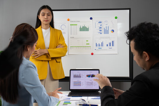 Asian businesswoman leading a meeting with her team, analyzing charts and graphs displayed on a whiteboard and laptop, demonstrating professionalism and collaboration in a corporate. - Powered by Adobe