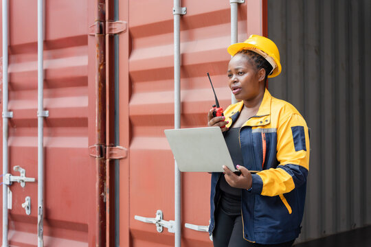 Confident dock worker managing logistics with laptop and radio at shipping terminal, Female industrial worker communicating via walkie talkie while using laptop at container yard - Powered by Adobe