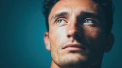 Fototapeta premium A close-up portrait of a young man with striking blue eyes, gazing thoughtfully upwards against a muted blue background.