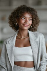A confident young woman with curly hair smiles softly, dressed in a chic gray suit and white top, exuding modern style and positivity in a bright indoor setting.