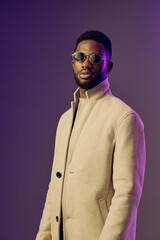 Stylish young Black man in a beige coat and sunglasses posing against a purple backdrop, exuding confidence and fashion forward vibes