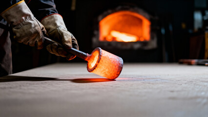 Gloved hands holding a red hot metal object near a burning furnace in a dark workshop for industrial production concept and traditional craftsmanship