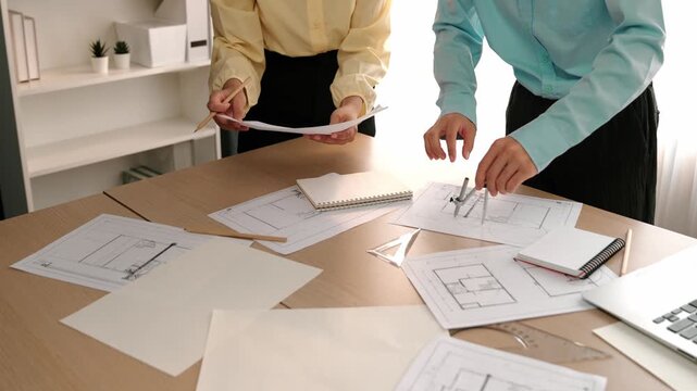 Architect and safety engineer discussion about building blueprint displaying on laptop on table with architectural document and wooden block scatter around at modern office. Closeup. Delineation.