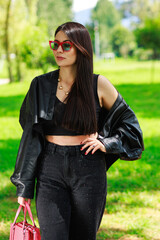 Young woman standing in a sunny park wearing sunglasses and holding a pink handbag