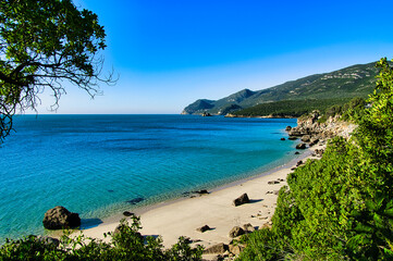 Serene coastal landscape with clear turquoise waters and lush green hills under a bright blue sky at the Atlantic coast near Sesimbra, Portugal
