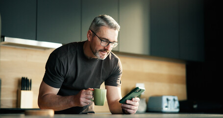 Man, coffee and scroll in kitchen with phone for social media notification, text message and news feed. Mature person, hot drink and browse with tech in home for online chat, reading article or blog.