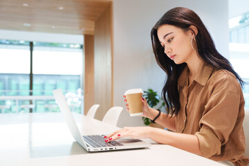 Young beautiful, stylish, elegant Asian woman in brown shirt hold coffee cup, work on computer laptop in luxury white office. Digital technology, modern lifestyle, online business and social media