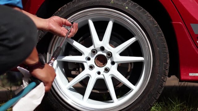 A close-up shot of a person using a tire inflator and pressure gauge on the valve stem of a red car's custom white and silver alloy wheel to check or add air pressure.