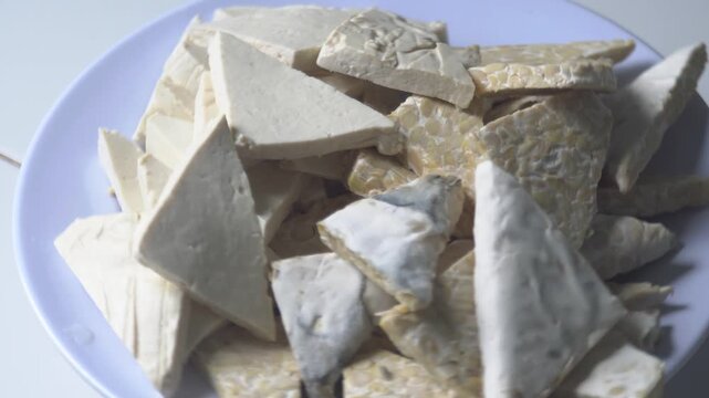 Top-down, static shot of a white plate filled with raw, triangular slices of tempeh and tofu. This combination of Indonesian high-protein ingredients is a cornerstone of vegan and vegetarian cooking