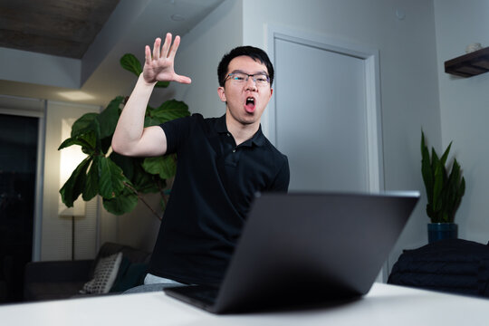 An Asian man wearing glasses makes a dramatic mic drop hand gesture while sitting at his desk with a laptop. He appears triumphant after finishing a task while working at night in his home office.
