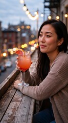 Fototapeta premium A young Asian woman relaxes with a pink fruit cocktail on an urban balcony. Vertical portrait of a person enjoying a drink in the evening with a city view and bokeh lights in the background