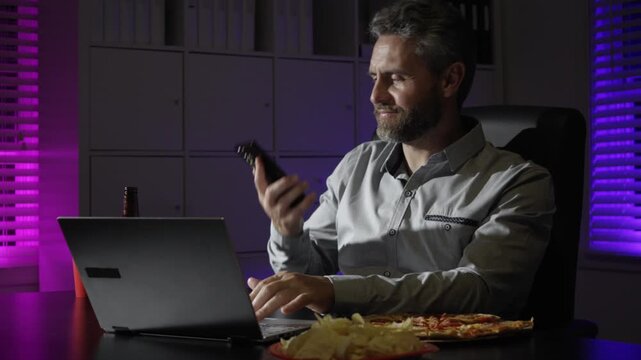 Freelancer working late with snacks in night home office. Business man night routine with laptop. Snack break during laptop work in office. Man at desk eating popcorn in late hours.