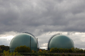 Two large spherical gas storage tanks stand against a dramatic cloudy sky, surrounded by greenery, showcasing industrial architecture and environmental integration