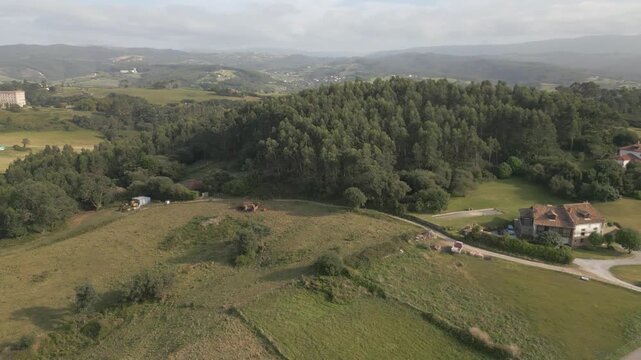 Stationary drone shot with a left yaw over the outer fields of Comillas, showing green meadows, tree-filled estates, the iconic university behind, and the coastline with its beaches.