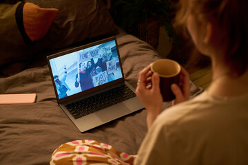 Young adult Caucasian woman sitting on bed holding mug, watching protest footage on laptop screen showing diverse group of people holding signs and demonstrating indoors