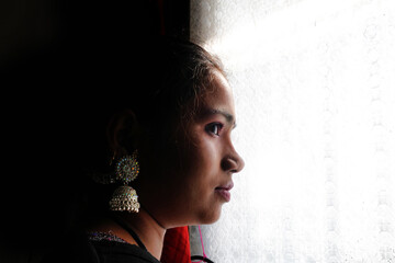 Side profile of an Indian woman looking through a window in dramatic natural light, wearing traditional earrings, thoughtful mood and quiet emotion, artistic lifestyle portrait with contrast shadow an