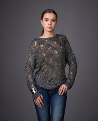 Young woman in a trendy shiny, open-knit sweater and jeans posing against a gray background. Casual glamour style.