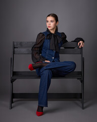 Studio fashion portrait of a young woman wearing a dark denim overalls or a set consisting of a structured vest and flared trousers, paired with a black sheer blouse, a tie and red high heels.