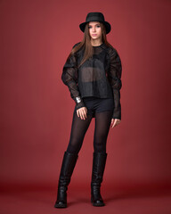 Dynamic studio fashion shot of a woman in a sheer black top, shorts, high boots, and fedora, walking against a vivid red background. Edgy, bold, and contemporary style.