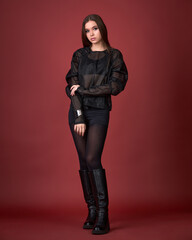 Dynamic studio fashion shot of a woman in a sheer black top, shorts, high boots, and fedora, walking against a vivid red background. Edgy, bold, and contemporary style.