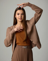 A studio portrait of a young woman posing playfully in a soft brown and beige ensemble consisting of a pleated midi skirt, a knit cardigan, and hoop earrings. Her arms are raised above her head.