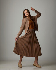 A studio fashion portrait of a young woman playfully posing in a monochrome brown outfit consisting of a pleated midi skirt, an off-the-shoulder knit cardigan, and loafers.