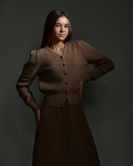 A close-up studio portrait of a young woman wearing a modest brown knit cardigan and pleated midi skirt, posing with her hands on her hips. Dramatic, subdued lighting against a dark background.