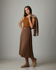 A studio fashion portrait of a young woman wearing a voluminous bright yellow poncho or turtleneck sweater cinched with a wide leather belt, large gold jewelry, and a brown felt hat.