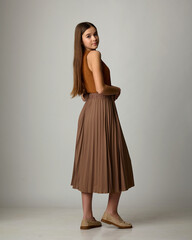 A studio fashion portrait of a young woman, seen from behind and in profile, wearing a monochrome brown ensemble consisting of a pleated midi skirt, a simple blouse, and loafers. Posing against a ligh