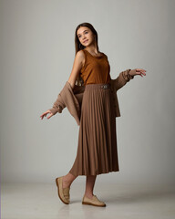 A studio fashion portrait of a young woman playfully posing in a monochrome brown outfit consisting of a pleated midi skirt, an off-the-shoulder knit cardigan, and loafers.