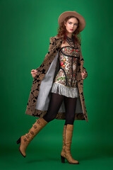 A full-length studio fashion portrait of a young woman against a bright green background. She is wearing a brown velvet coat with a floral print, black tights, and tall brown leather boots.