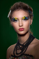 A striking close-up portrait of a young woman, adorned with intricate, colorful floral and pearl elements around her eyes. The high-fashion, fantasy makeup creates a vibrant, artistic image.