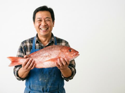 Smiling Asian man in plaid shirt and blue apron holding a large fresh red snapper fish against a plain white background