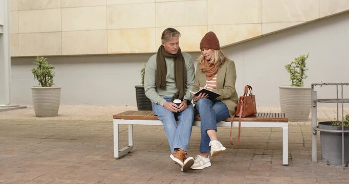 Senior couple turning book pages on bench in plaza, woman reading aloud while man leaning closer - Powered by Adobe