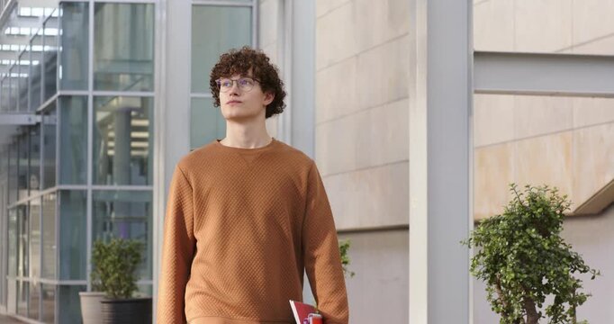 Entering frame, student carrying folder walking down outdoor corridor, examining building design
