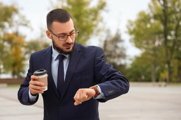 Young male businessman with coffee looking at wristwatch outdoors. Time management concept
