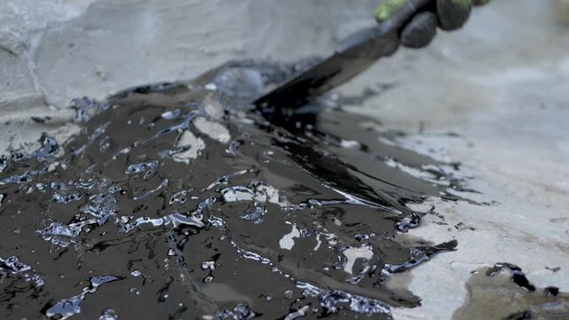 Bituminous waterproofing putty is being applied to rough sand-cement surface with trowel by gloved hand. Putty is sticky viscous and waterproof used to safeguard buildings from moisture. Seal cracks