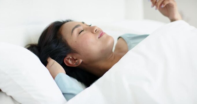 Mid adult Asian female gazing away, pushing aside duvet, stretching arms overhead, savoring morning
