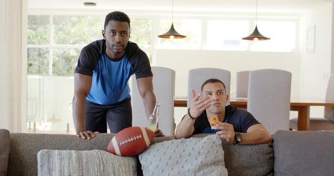 Diverse male friends leaning on couch crunching chicken reacting to intense game with football