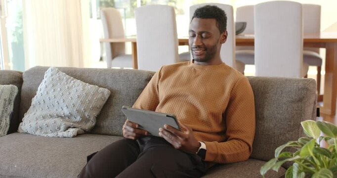 African American man entering living room sitting on gray couch tapping tablet browsing articles