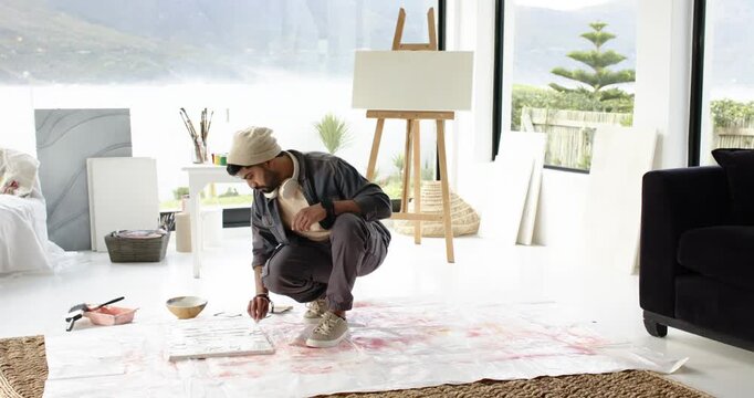 Indian man picking paintbrush from bowl and painting block pattern on floor canvas in studio