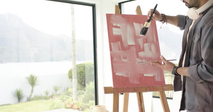Artist dipping brush and applying crossing white strokes on red canvas at window forming grid