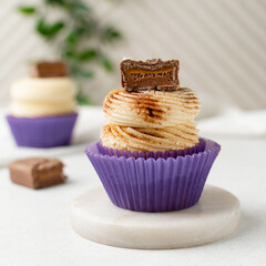 Close-up of a vanilla cupcake in a purple wrapper with buttercream frosting, cinnamon dust, and a caramel-filled chocolate, cupcake in a purple liner with a cinnamon frosting and caramel chocolate bar