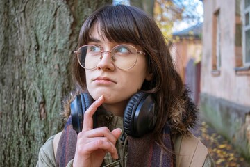 Emotion of thoughtfulness and dreaminess on the face of a young woman in glasses with headphones, outdoors