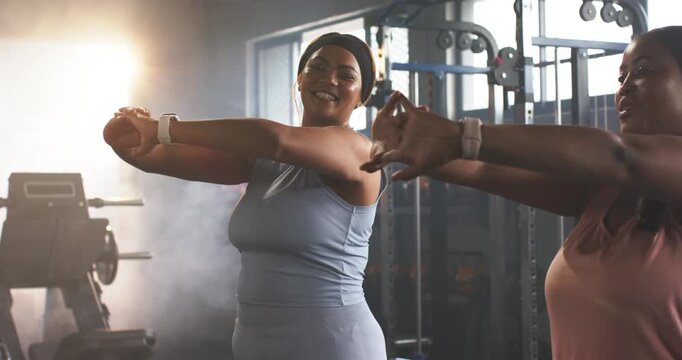 African American women in tanks following right woman's cue, warming arm stretches by gym cable
