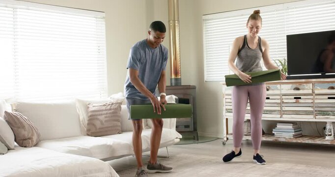 Diverse couple entering living room in activewear holding green yoga mats, unrolling for workout