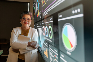 An Asian businesswoman stands confidently in a modern office, holding a tablet beside large digital...