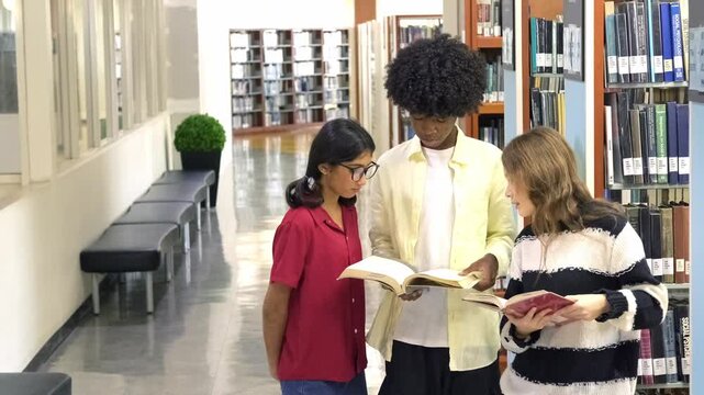 Three cheerful students stand in a bright library hallway, smiling and comparing books beside tall shelves, symbolizing youth education, learning resources, and academic engagement.