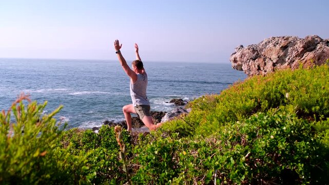 Fit man does yoga warrior poses on coastal cliff overlooking ocean at sunrise