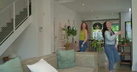 Two female friends greeting, opening arms, dancing across bright home sofa, denim jacket green top